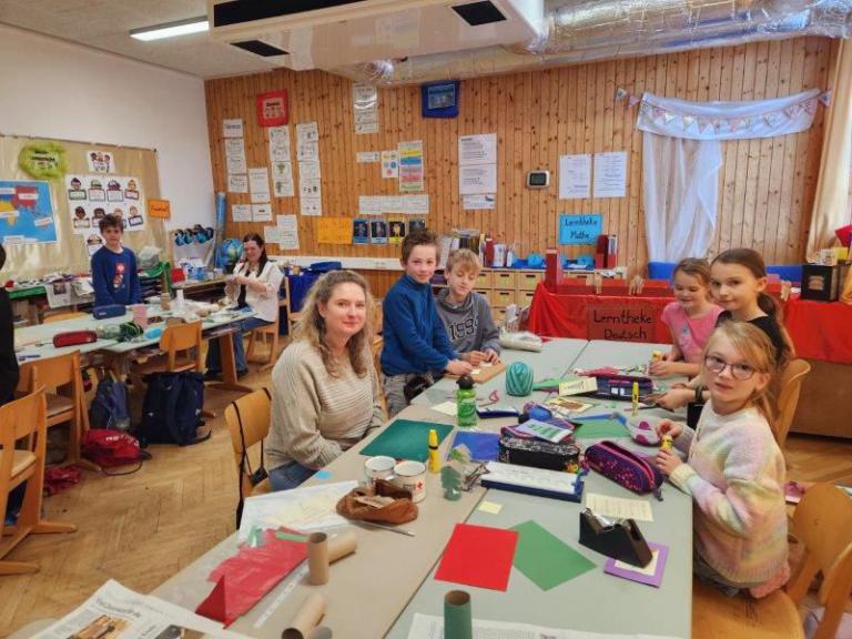 Kinder und Erwachsene sitzen an Tischen in einem Klassenraum und basteln mit Papier, Kleber und Scheren.