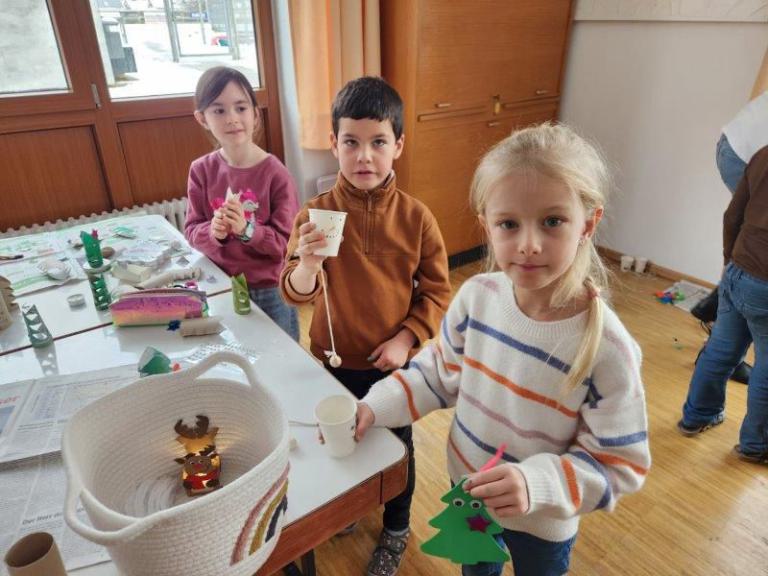 Drei Kinder sitzen an einem Tisch mit Bastelmaterialien, eines hält einen grünen Papier-Weihnachtsbaum