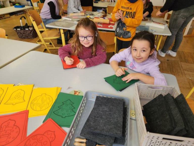 Zwei Kinder sitzen an einem Tisch und basteln mit buntem Papier, darunter grüne und rote Blätter mit Weihnachtsmotiven