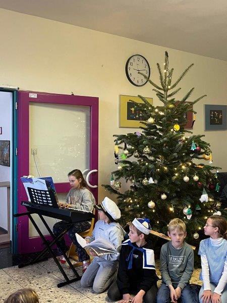 Mehrere Kinder vor einem geschmückten Weihnachtsbaum, ein Kind spielt Keyboard, im Hintergrund eine Uhr an der Wand.