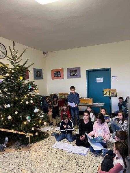Weihnachtsbaum mit Kugeln und Lichterkette, mehrere Kinder sitzen und stehen im Raum, einige halten Notenblätter.