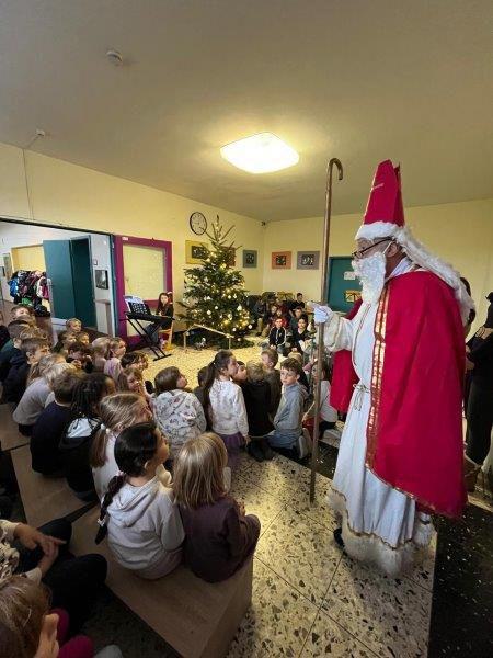 Weihnachtsmann mit rotem Mantel und Bischofsstab steht vor einer Gruppe sitzender Kinder or einem Weihnachtsbaum.