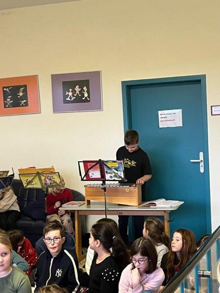 Mehrere Kinder sitzen auf dem Boden, eine Person steht hinter einem Tisch mit einem Musikinstrument, im Hintergrund eine blaue Tür mit einem Schild.