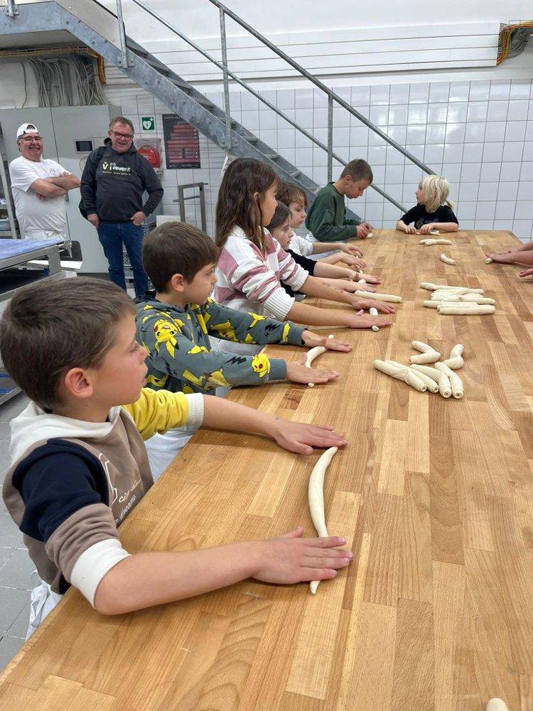 Mehrere Kinder sitzen an einem langen Holztisch und formen längliche Teigstücke, im Hintergrund stehen zwei Erwachsene vor einer Treppe.