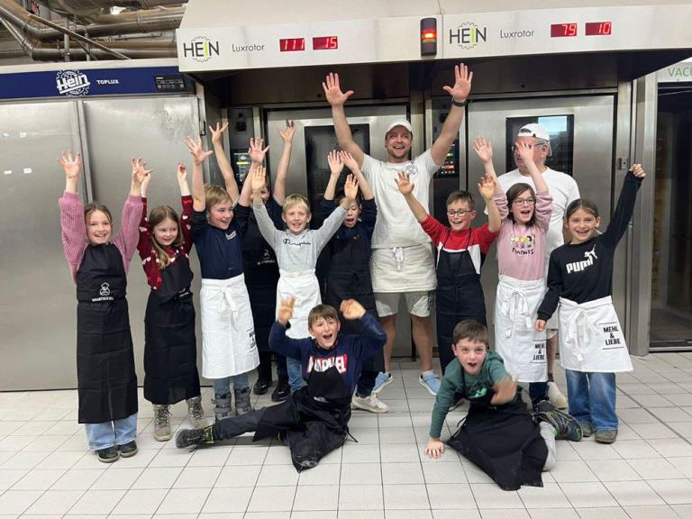 Kinder und zwei Bäcker stehen vor großen Bäckereiöfen und heben die Hände hoch