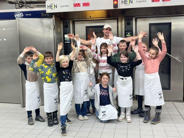 Gruppe von Kindern und einem Erwachsenen in einer Bäckerei, alle tragen weiße Schürzen und heben die Hände.