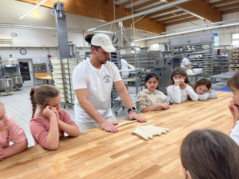 Bäcker in weißem T-Shirt und Mütze zeigt Kindern an einem Holztisch mehrere längliche Teigstücke in einer Bäckerei.