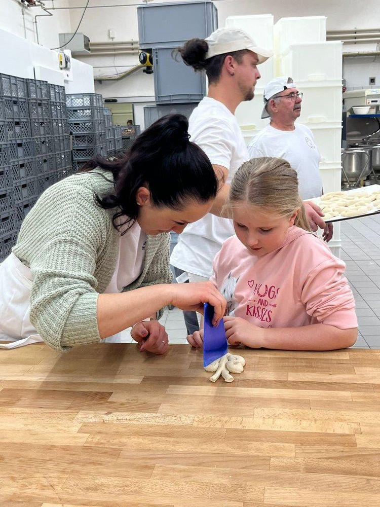 Frau und Mädchen formen gemeinsam Teigstücke auf einem Holztisch in einer Bäckerei, im Hintergrund zwei Personen in weißer Arbeitskleidung.