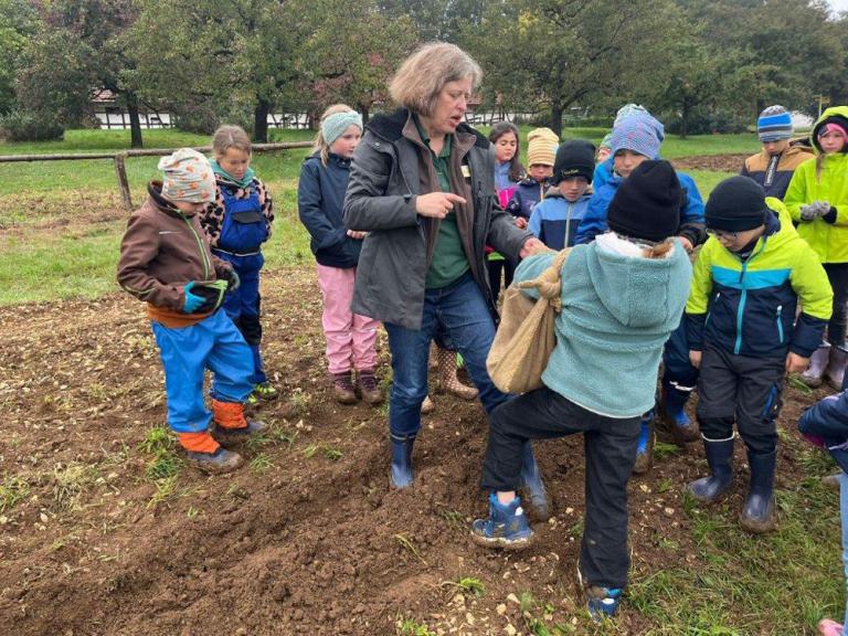 Gruppe von Kindern mit bunter Outdoor-Kleidung und Gummistiefeln steht auf einem Acker, eine Frau in schwarzer Jacke zeigt auf den Boden.
