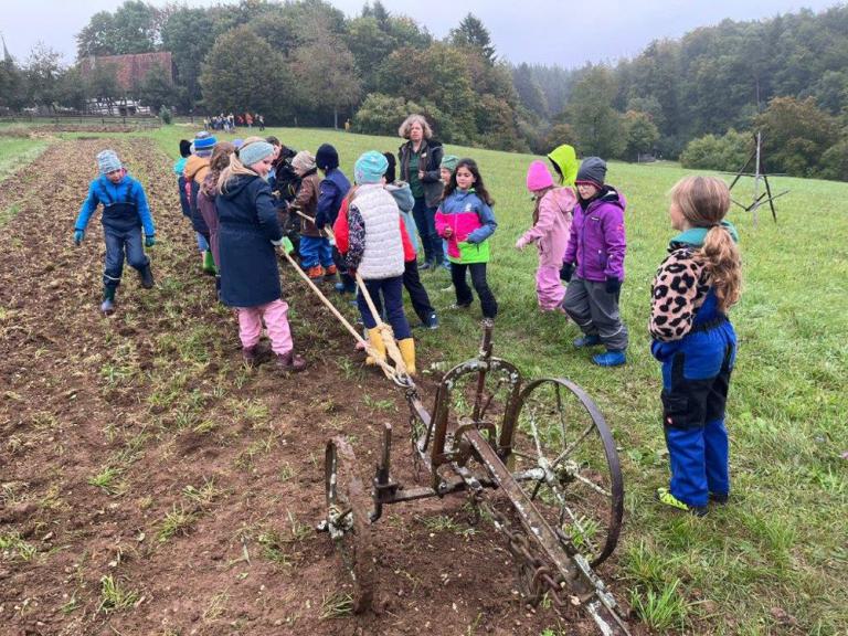 Gruppe von Kindern in bunter Outdoor-Kleidung steht auf einem Feld mit einem alten Pflug