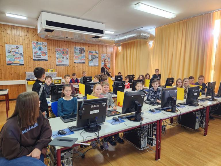 Klassenraum mit mehreren Kindern an Computertischen, eine Lehrerin steht vorne