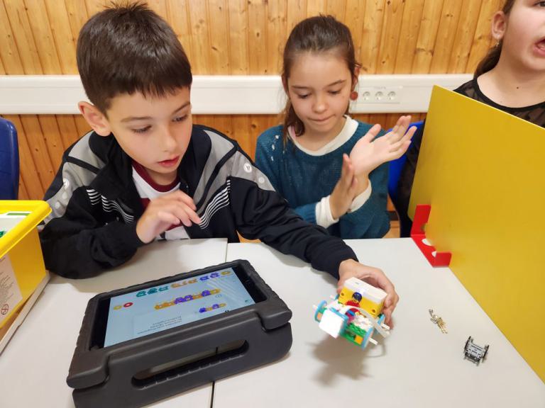 Zwei Kinder sitzen an einem Tisch, eines hält einen kleinen Roboter aus bunten Bausteinen, davor liegt ein Tablet in einer schwarzen Schutzhülle.