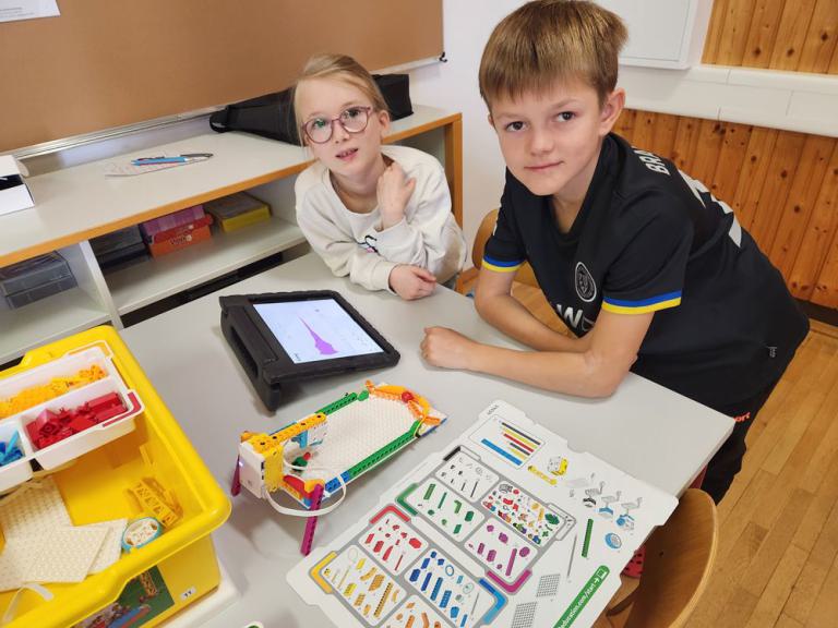 Zwei Jungen an einem Tisch mit Tablet, Baukasten mit bunten Bauteilen und Anleitung für ein Roboterprojekt.