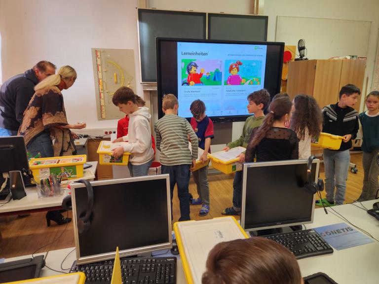Kinder stehen in einem Klassenraum und halten gelbe Kisten, im Hintergrund ein Bildschirm mit Lerninhalten und Erwachsene, die helfen.