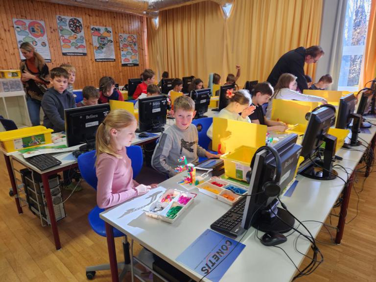 Mehrere Kinder sitzen an Computertischen in einem Klassenraum, einige arbeiten mit bunten Bausteinen und gelben Trennwänden.