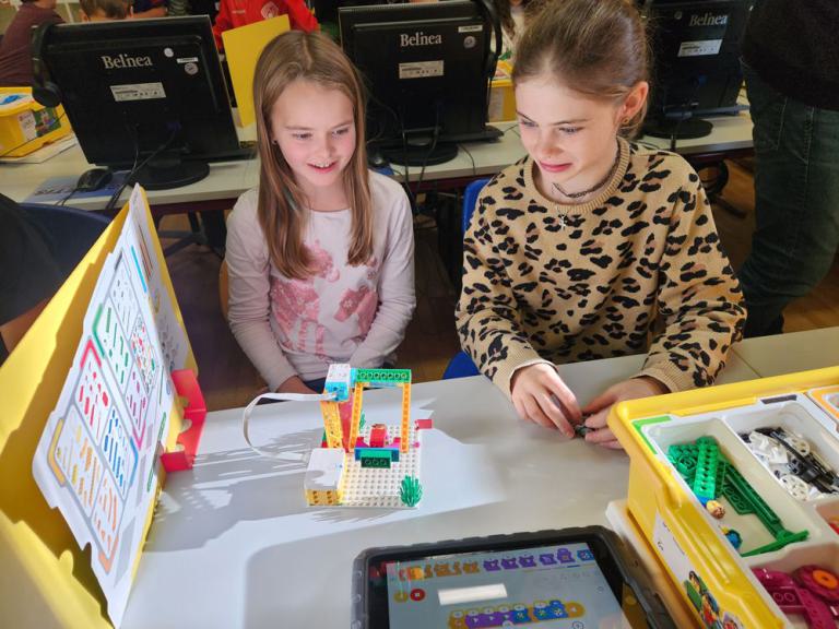 Zwei Mädchen sitzen an einem Tisch mit einem Lego-Roboter, einem Tablet mit Programmieroberfläche und einer Anleitung auf einem Ständer.