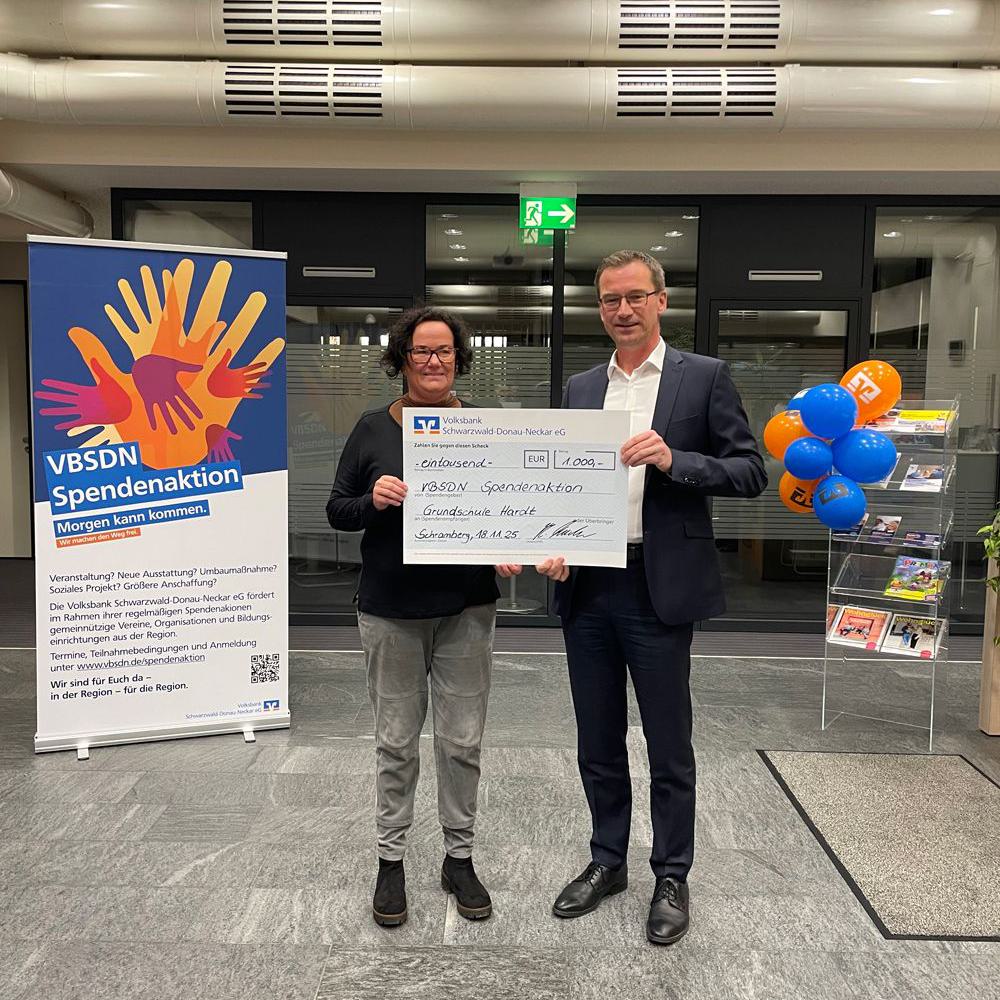 Zwei Personen stehen nebeneinander und halten einen großen Spendenscheck in einem Innenraum mit Informationsstand und Luftballons.