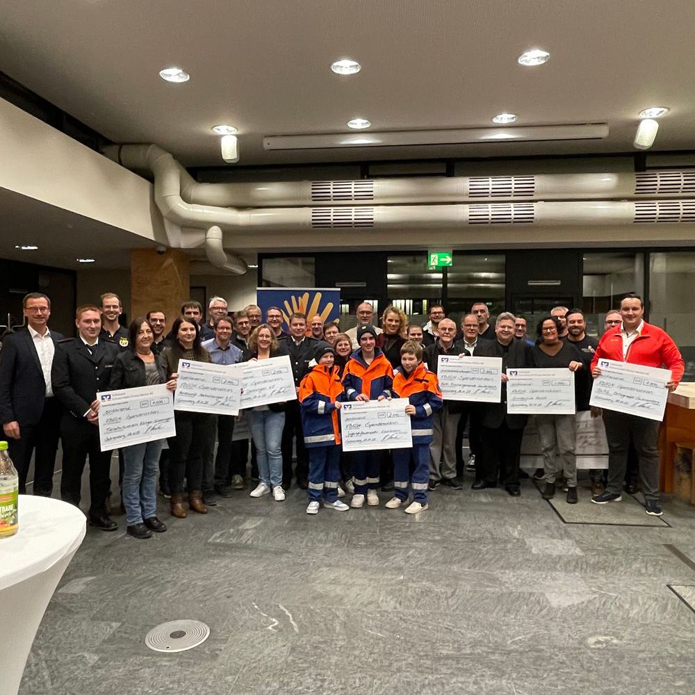 Große Gruppe von Menschen in einem Raum, einige tragen orangeblaue Arbeitskleidung, mehrere halten große Spendenschecks.