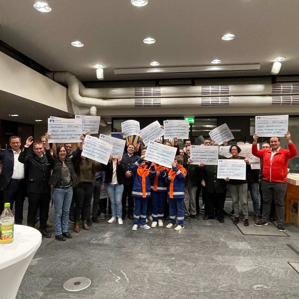 Gruppe von Menschen in Innenraum, einige Kinder in orange-blauer Arbeitskleidung, halten große Spendenchecks hoch.