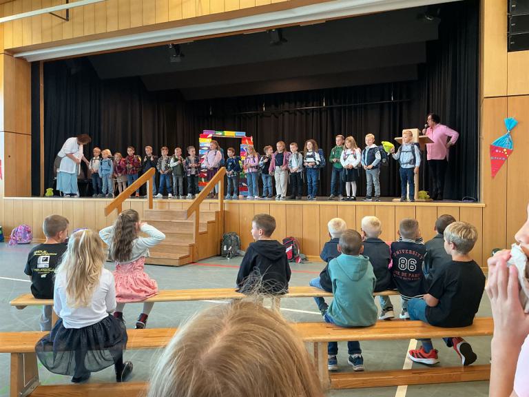 Eine Gruppe von Kindern sitzt und steht in einem großen Saal, während auf der Bühne eine Aufführung stattfindet.