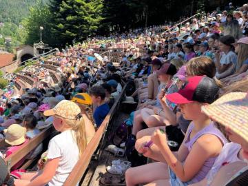 Blick über ein voll besetztes Freilufttheater, in dem vor allem Kinder mit Sonnenhüten dicht an dicht auf Holzbänken sitzen.