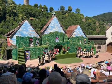 Schauspieler in Kostümen auf einer grünen Freilichtbühne mit Kulissenhäusern vor Publikum unter blauem Himmel.