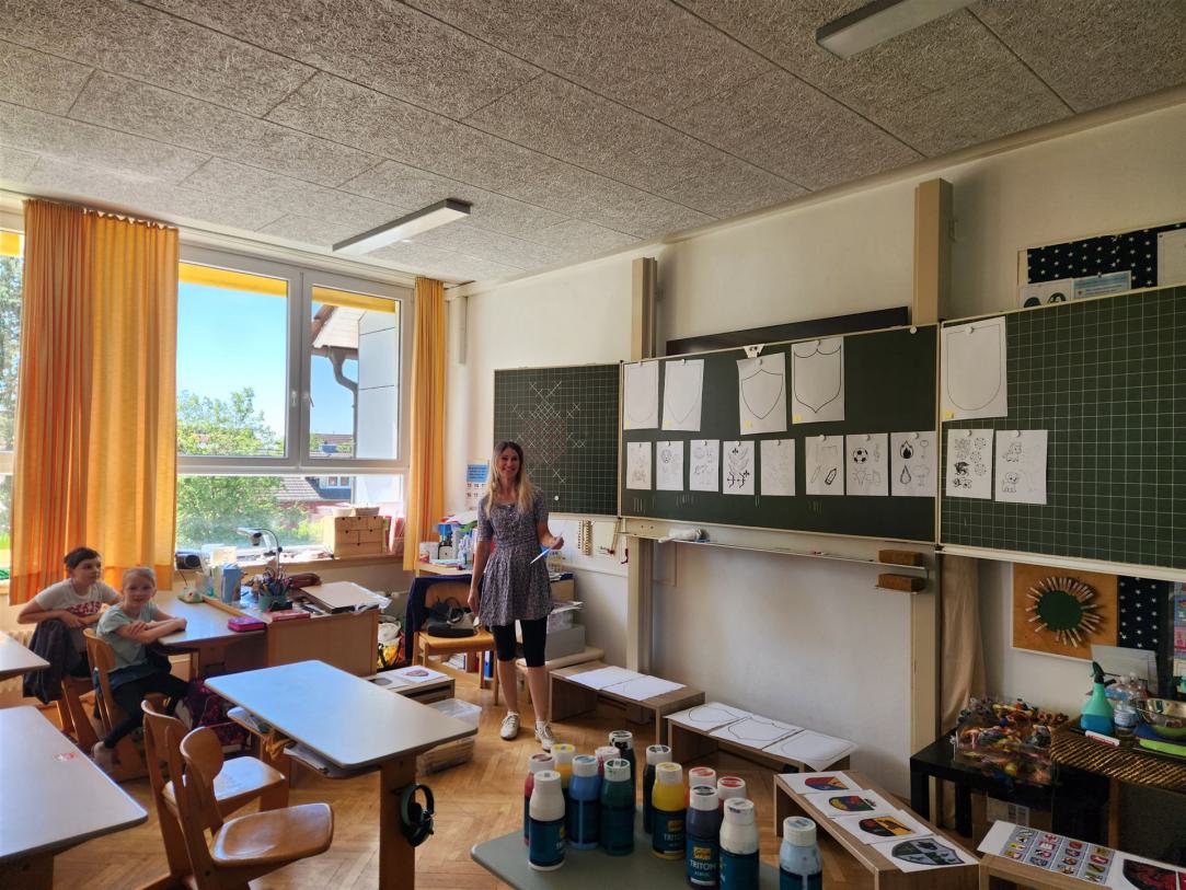 Das Innere eines hellen und großen Klassenzimmers mit großen Fenstern, zahlreichen Tischen und einer Präsentationswand voller Lernmaterialien.