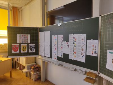 Eine große Tafel in einem Klassenzimmer, die mit Lernmaterialien über Wappenkunde und Heroldsmerkmale bedeckt ist.