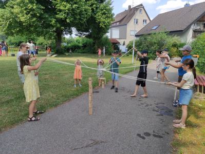 Kinder und Erwachsene spielen ein Seilspiel auf einem asphaltierten Weg in einer Grünanlage vor Wohnhäusern.